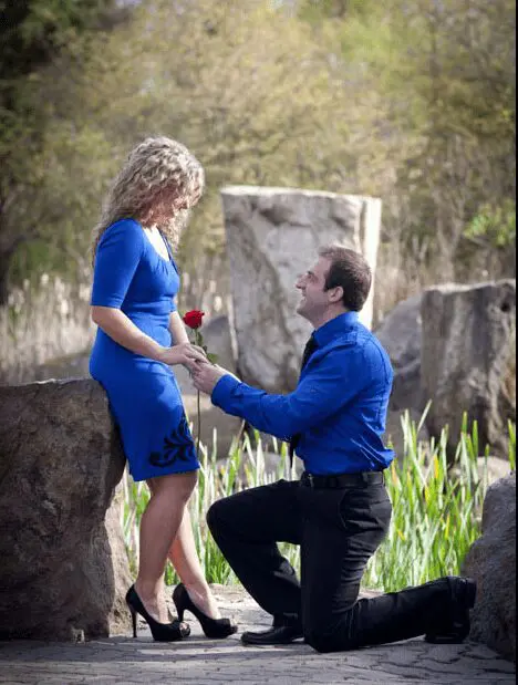 Guy in a blue long-sleeved formal shirt kneeling to offer a rose to his blond female partner